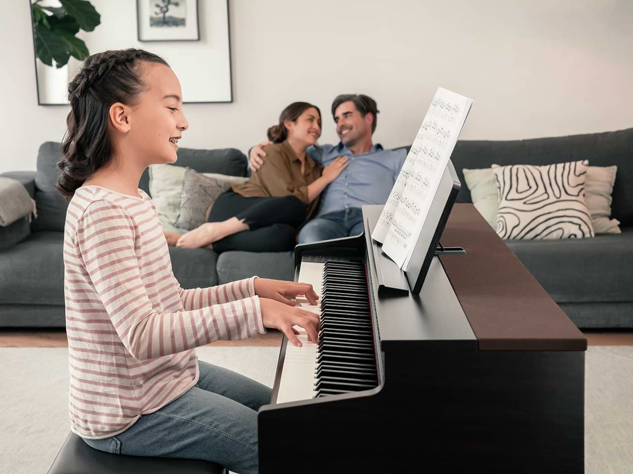 hình ảnh một bé gái đang chơi đàn piano Kawai CN 301 R tại nhà. Cũng trong căn phòng đó, bố mẹ cô gái đang ngồi nói chuyện với nhau
